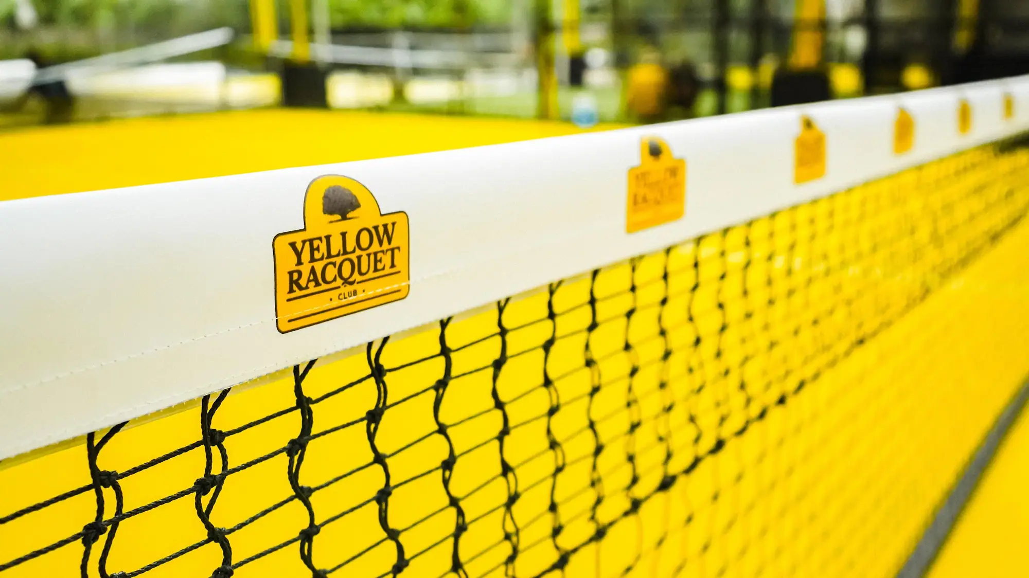 Yellow Racquet Club branded net over a yellow padel court